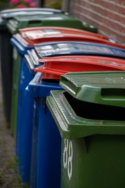 A large collection of mixed domestic waste and rubbish piled around a public waste collection point on a paved sidewalk. The pile includes various black and colored bin bags, flattened cardboard boxes