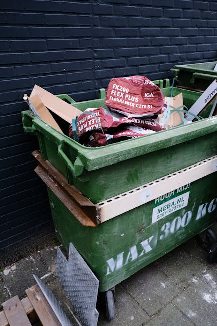 A green plastic waste bin positioned on a paved surface against a dark brick wall, filled with bags of garden or building materials. The bags are red and white with printed labels indicating their con